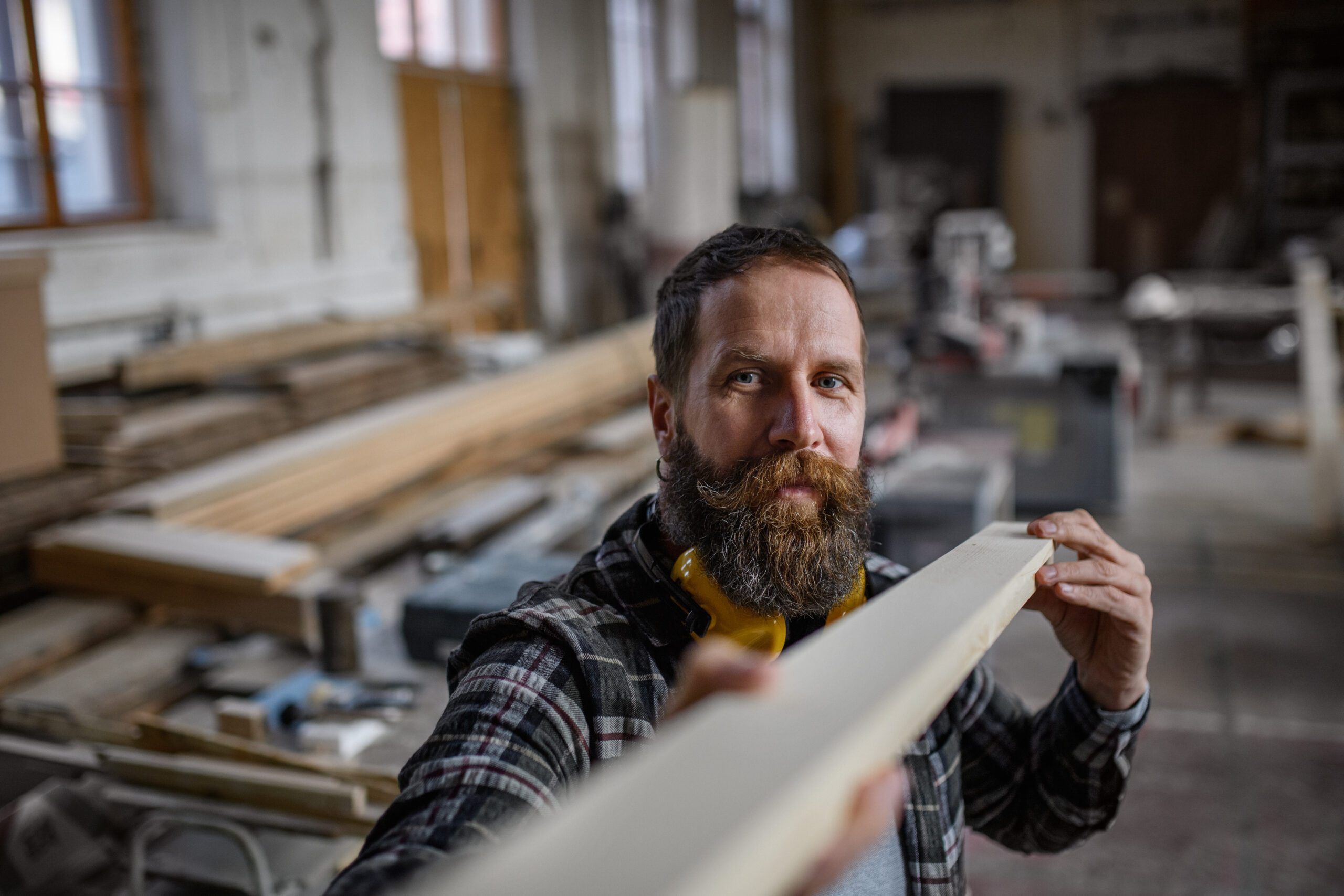 Mature male carpenter carrying wooden board indoors in carpentery workshop. Small business concept.